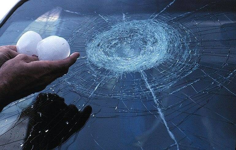 A hand holds two hailstones beside a windshield with a large circular fracture and radiating cracks.