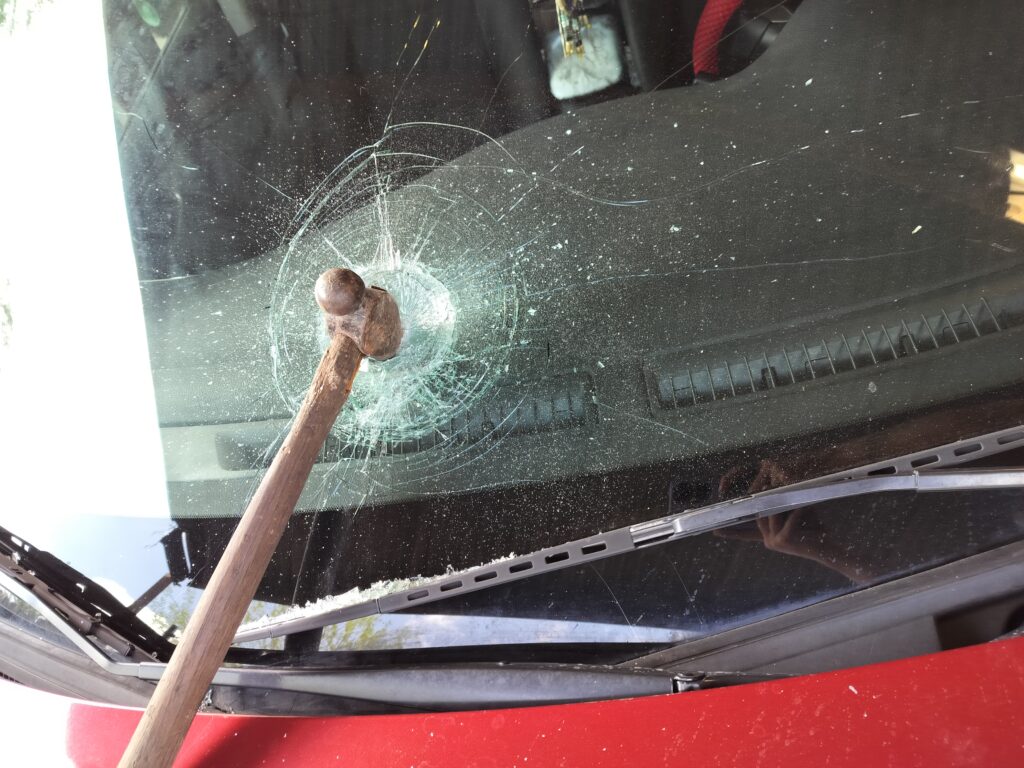 A wooden-handled ball-peen hammer lodged in a windshield, surrounded by circular impact cracks.